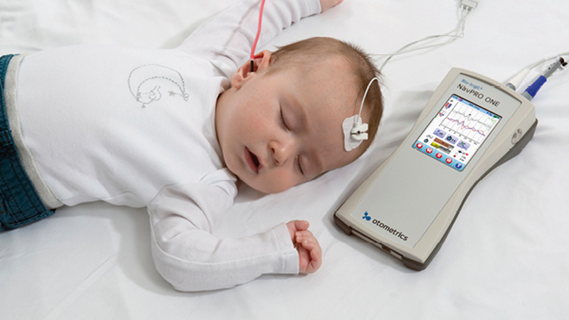 Baby prepared for pediatric ABR testing using a handheld auditory brainstem response device during natural sleep.