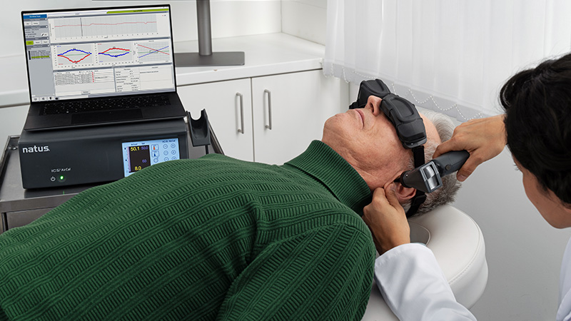 Clinician performing caloric irrigation with the Natus Sensory ICS Aircal on a patient wearing VNG goggles, with test results of diagnosis of balance disorders displayed on a connected laptop.