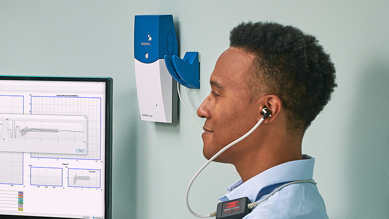 Adult undergoing OAE test with Madsen Capella2 device and otoacoustic emission response displayed on screen.